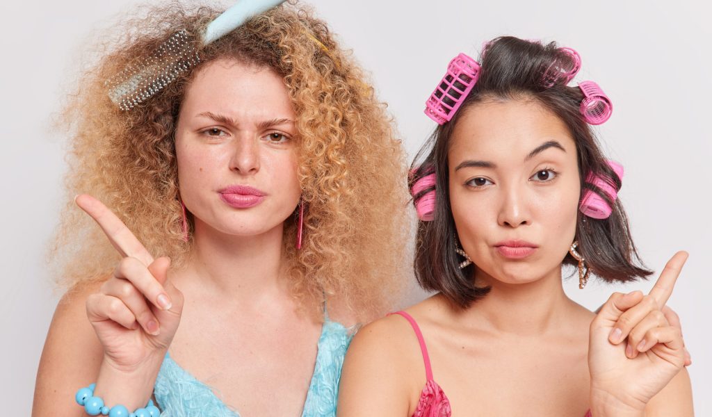 Serious women shake fingers say no look attentively at camera comb hair and apply rollers make hairstyle stand closely to each other get ready for special occasion isolated over white background